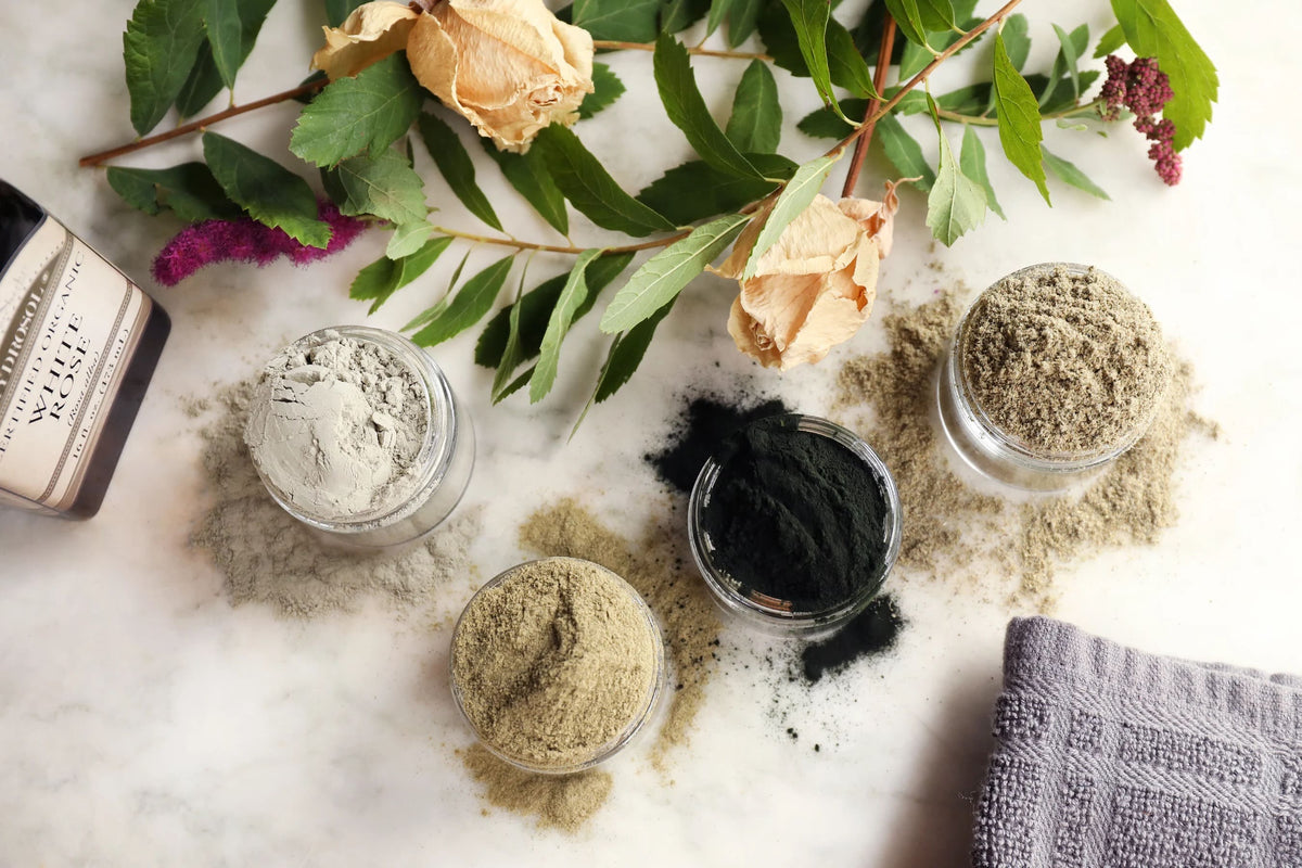 Four small clear jars filled with different colored powders arranged on a white marble surface with dried flowers, green leaves, and a gray cloth. A partial bottle label is visible at the left edge.