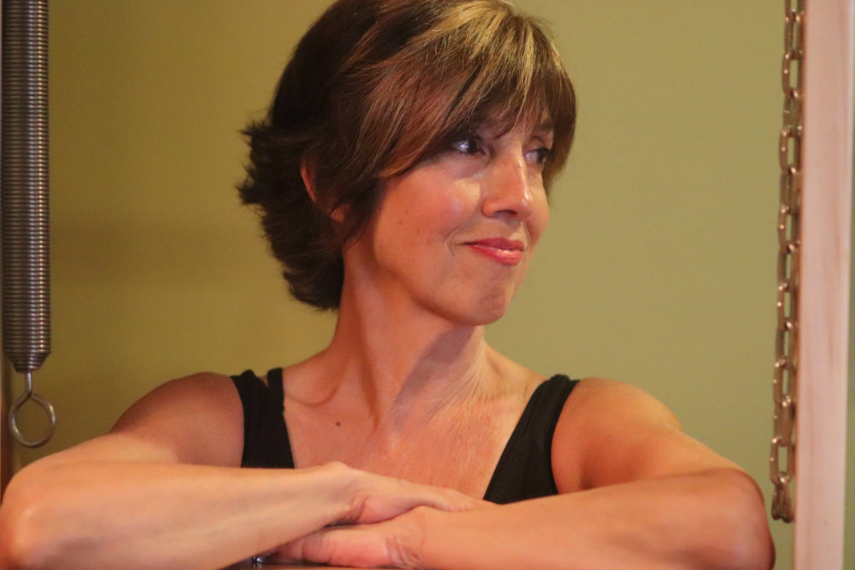 A woman with short brown hair wearing a black tank top, posing with her arms crossed in front of a light olive green wall. A metal spring and chain are visible on either side of the frame.