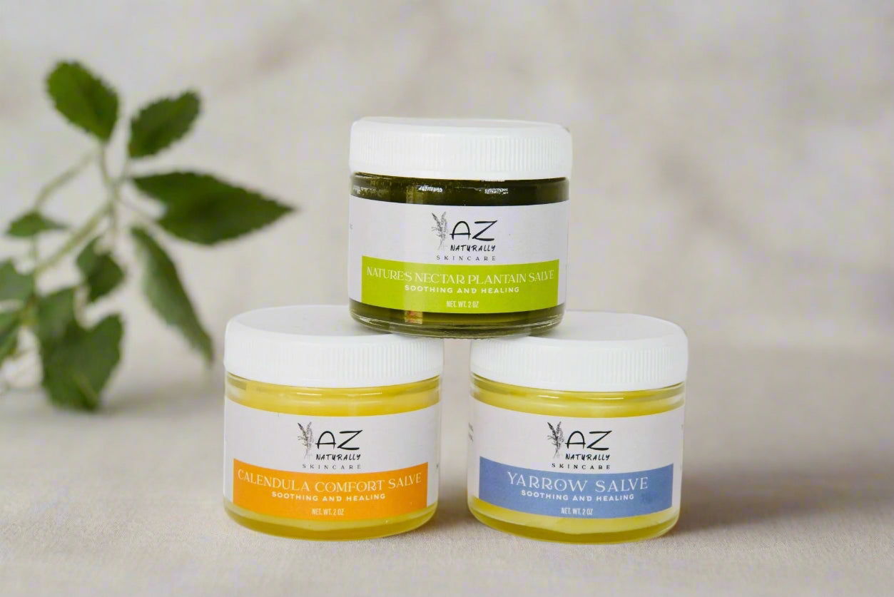 Three jars of herbal salve labeled 'Plaintain Salve', 'Calendula Salve', and 'Yarrow Salve' on a light gray surface with a leaf in the background