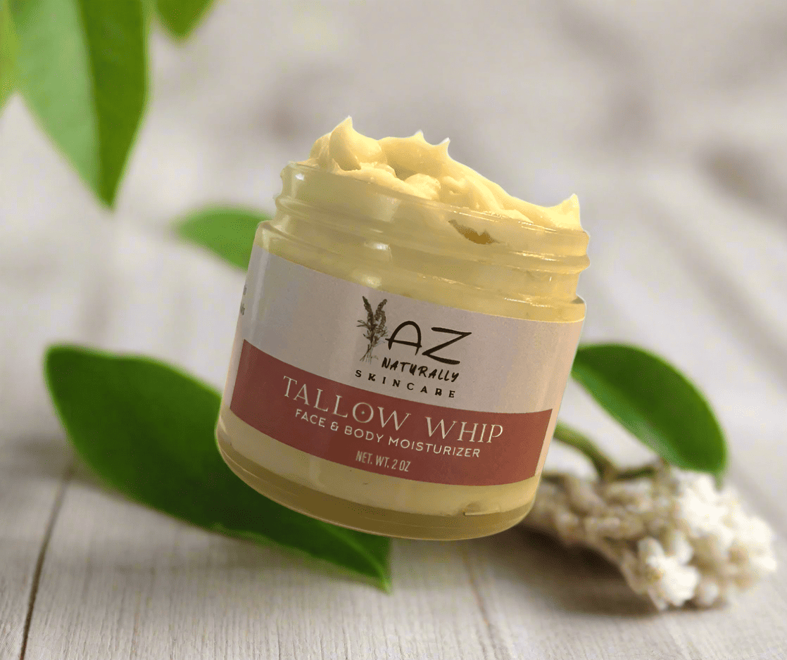 A jar of AZ Naturally Skincare Tallow Whip face and body moisturizer, filled with pale yellow cream and set on a light wooden surface with green leaves and small white flowers.