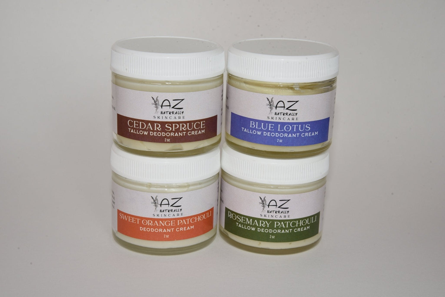 Four jars of 'Tallow Deodorant Cream' labeled 'Cedar Spruce', 'Blue Lotus', 'Sweet Orange Patchouli', and 'Rosemary Patchouli' on a light gray background
