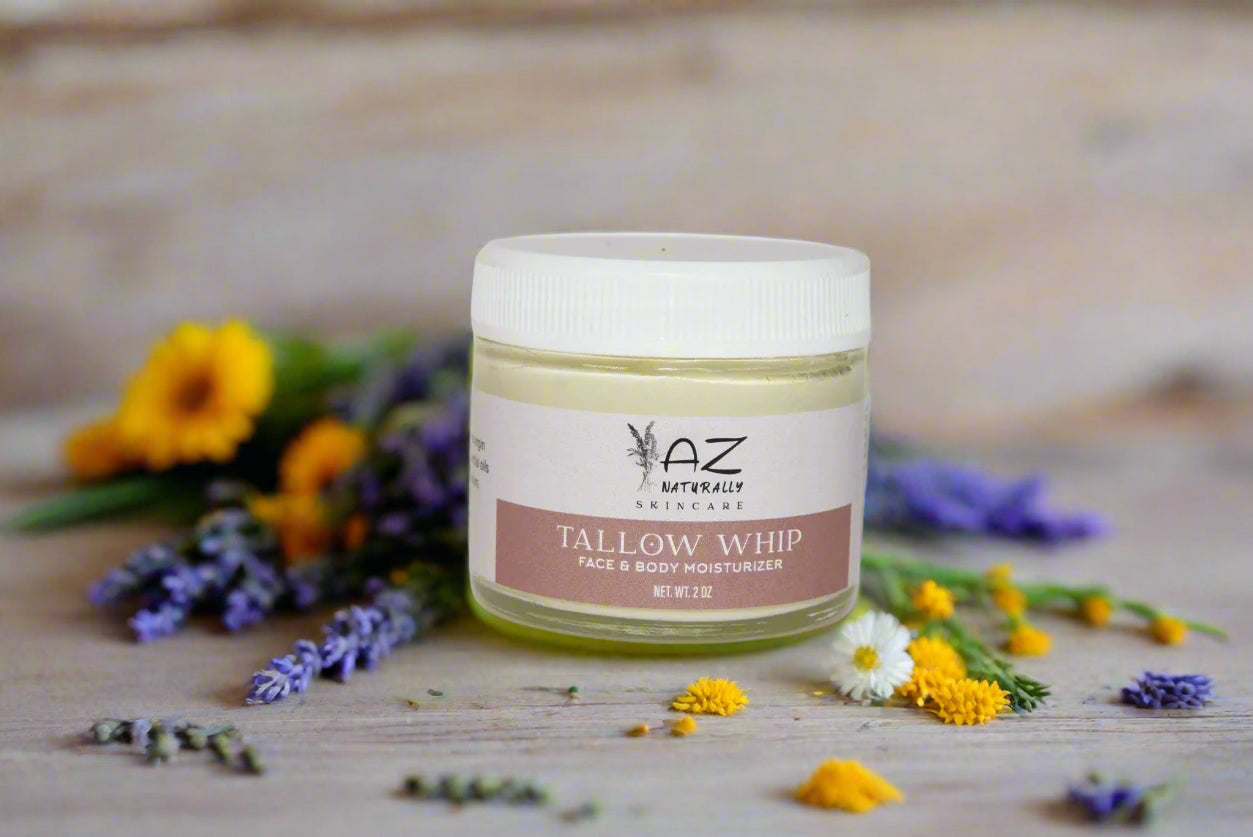 Jar of 'Tallow Whip Face & Body Moisturizer' surrounded by blue, white, and yellow flowers on a wooden surface 